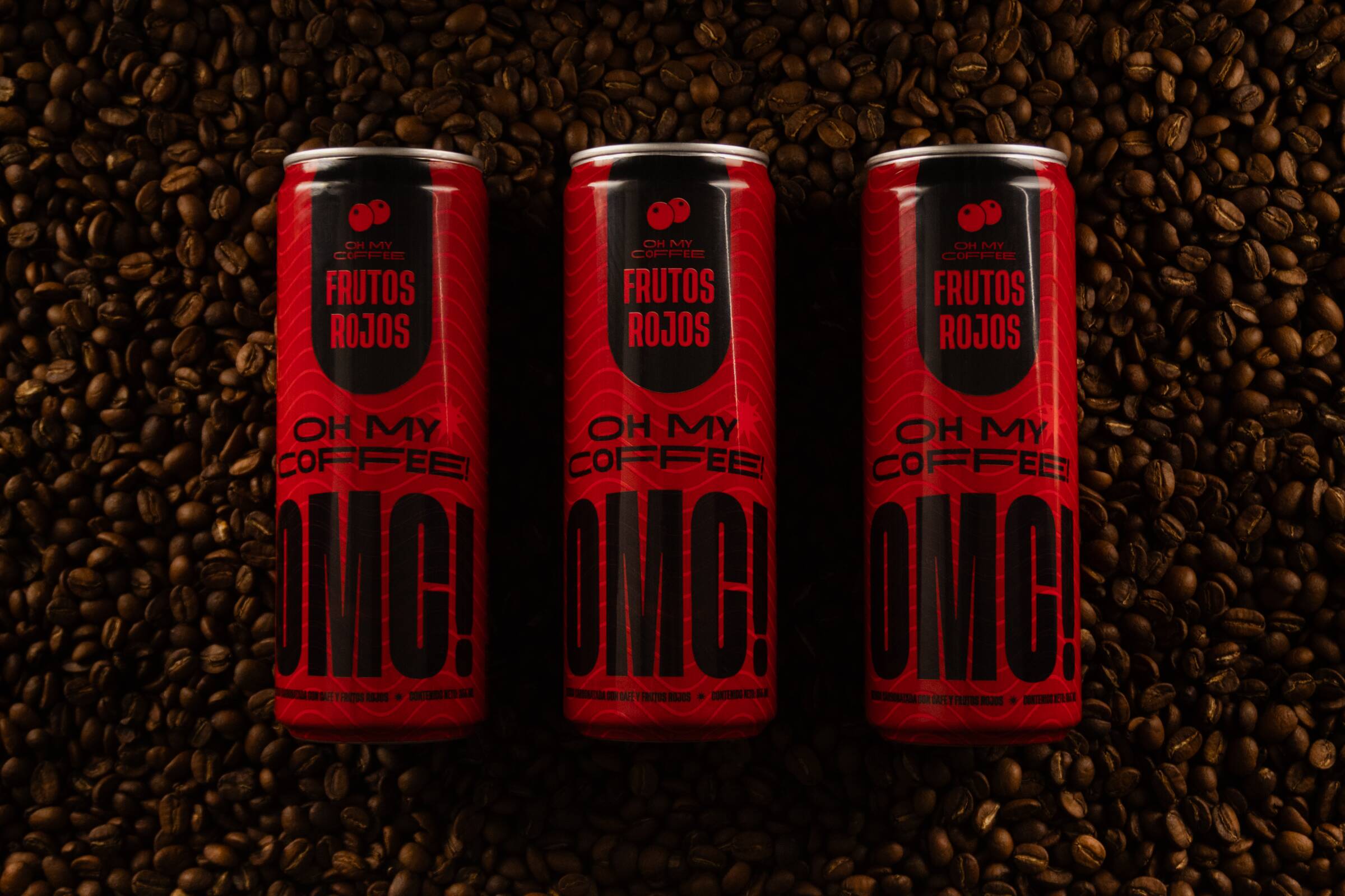 Tres latas de OMC! Frutos Rojos en composición cenital sobre fondo negro mate con granos de café